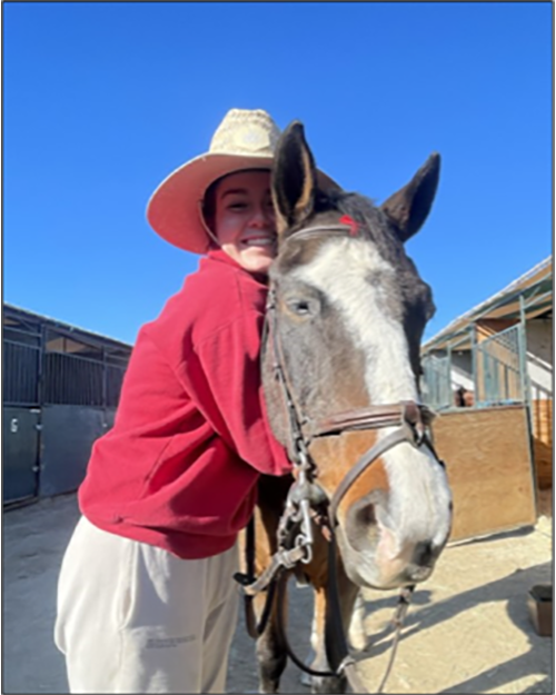 A smiling person wearing a cowboy hat, red long sleever shirt is hugging her horse around its neck.