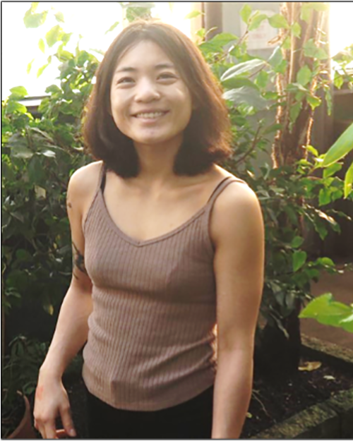 A smiling person with medium length brown hair wearing a brown tank top standing in from of a tall green plant
