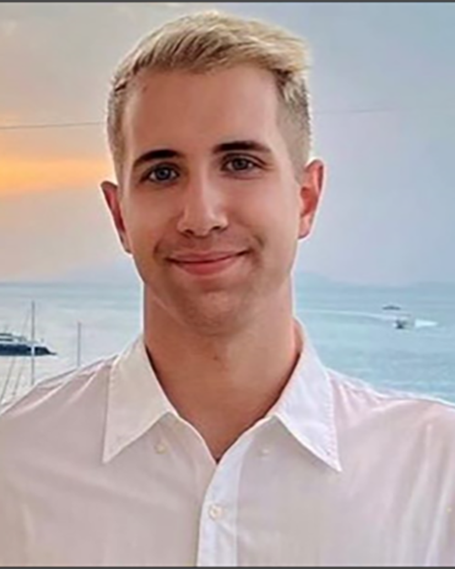 A smiling person presenting as a male with blond hair, white collared shirt. There is a sunset and ocean behind him.