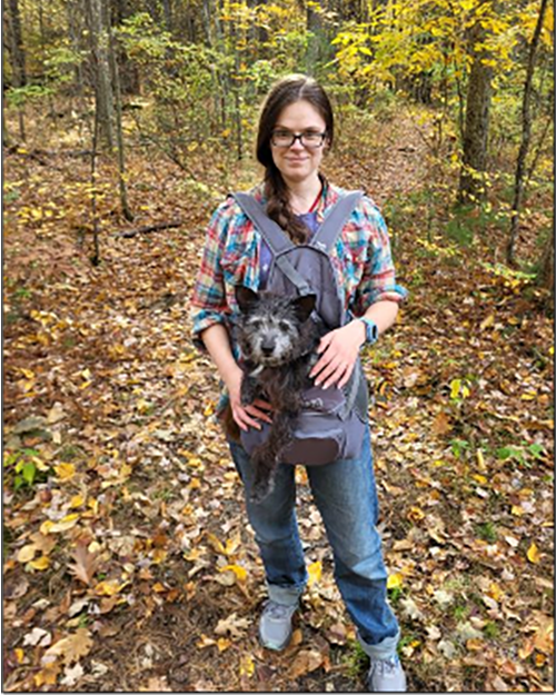 A person with long brown hair wearing eye glasses and carrying a dog in a backpack. Surrounded by fall leaves and trees.